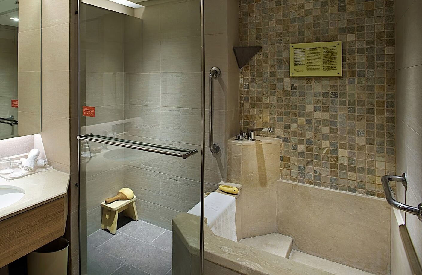 A modern bathroom with a glass shower enclosure, a bathtub with a mosaic tile wall, and a small stool.