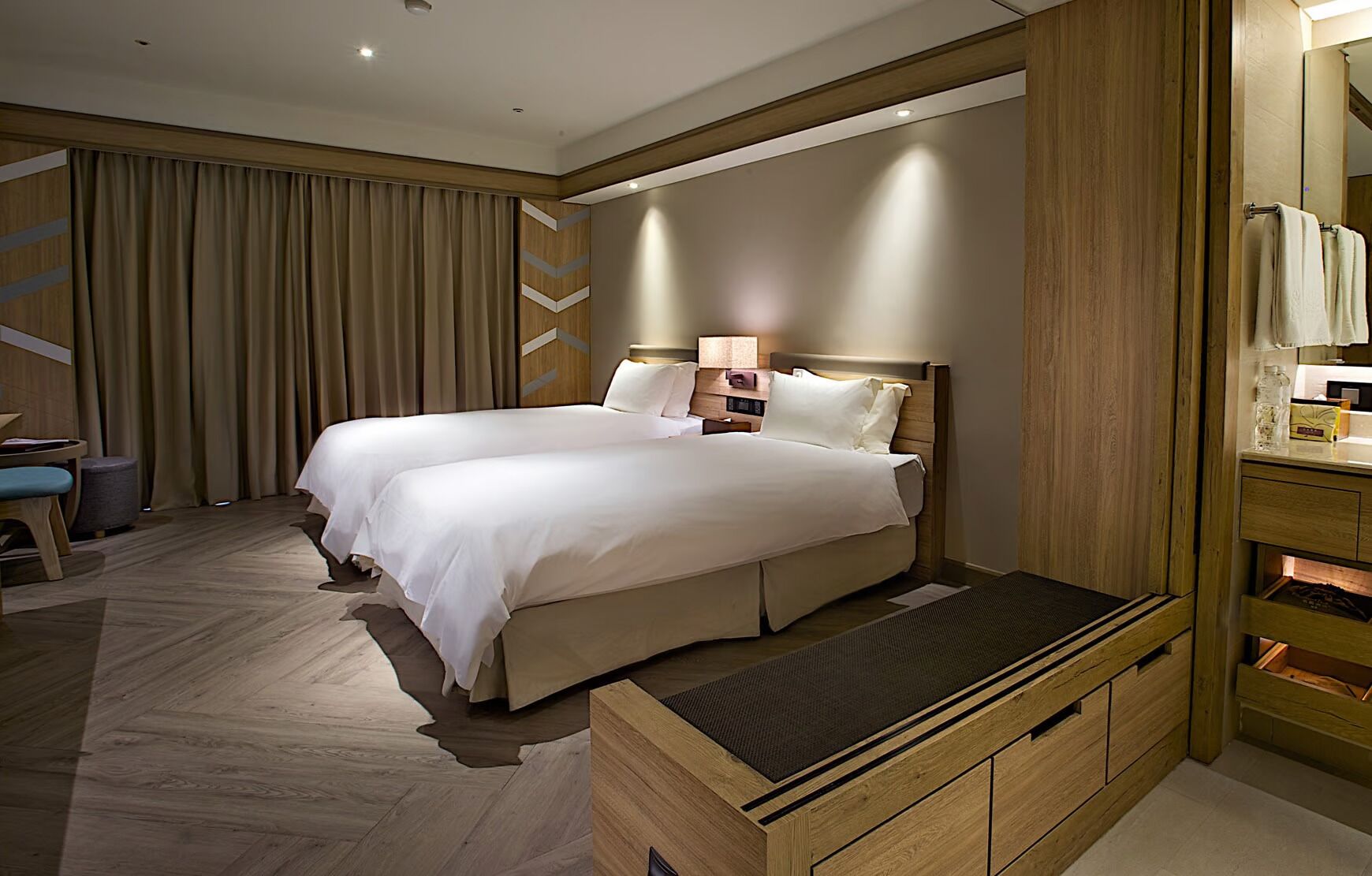 A hotel room with two beds, a wooden bench, a desk with a chair, and a large mirror.