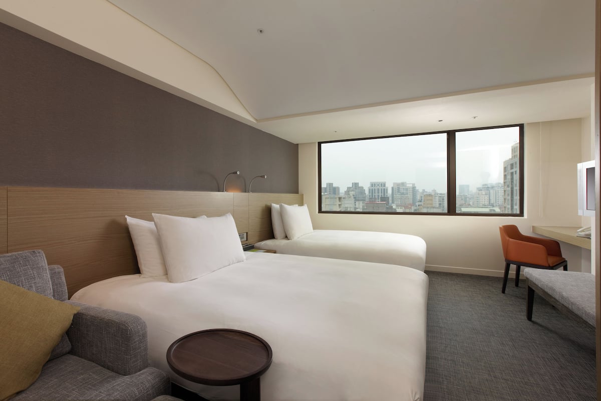 A hotel room with two beds, a sofa, a chair, a small table, and a large window with a city view.