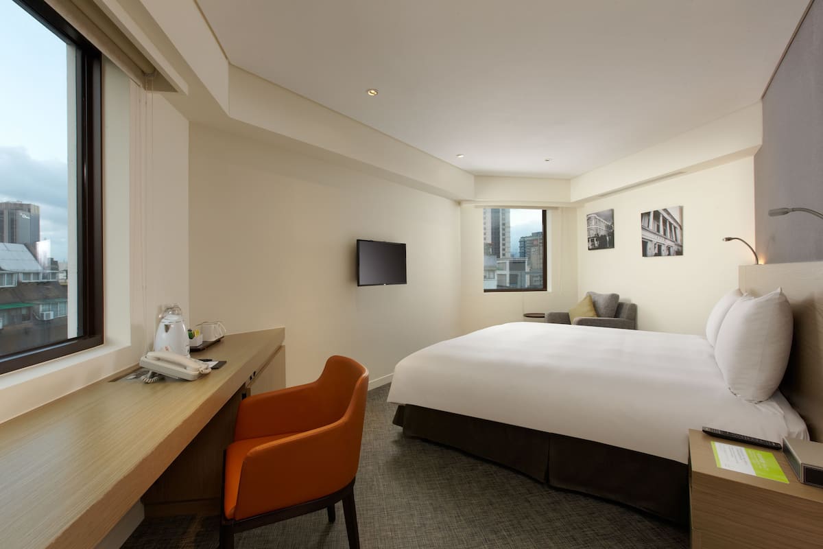 A hotel room with a bed, a desk, an orange chair, a TV, and a window with a city view.