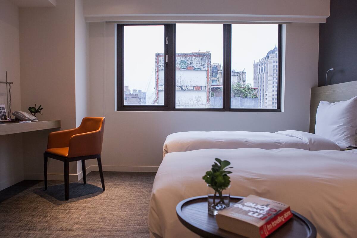 A hotel room with a bed, a desk with a chair, a window with a city view, and a small table with a book and a plant.