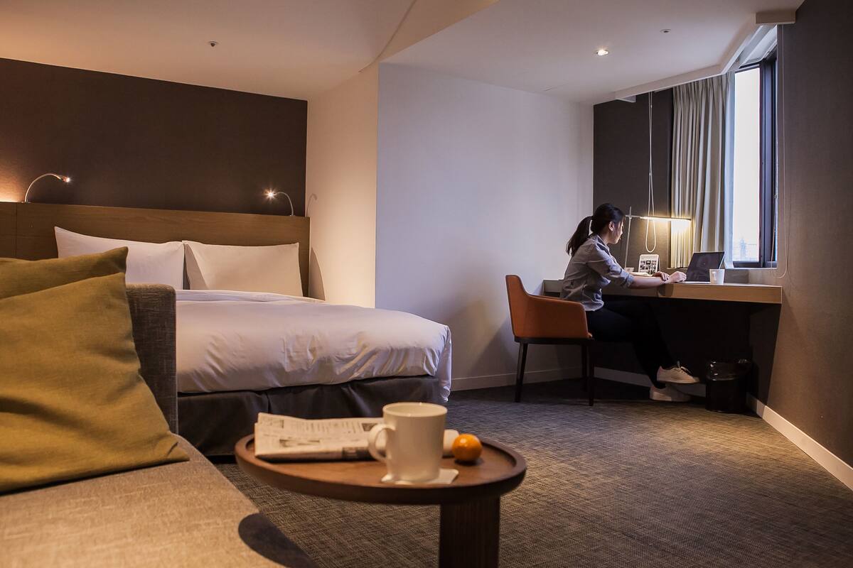 A hotel room with a bed, a desk, a chair, a coffee table with a cup and a book, and a window with curtains.