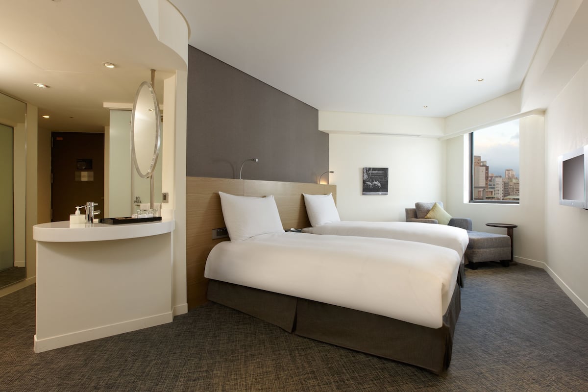 A hotel room with two beds, a vanity with a mirror, a small table, and a window with a city view.
