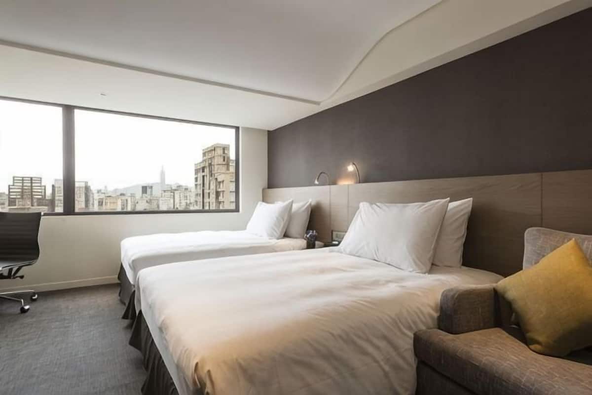 Ein Hotelzimmer mit zwei Betten, einem Sofa, einem Schreibtisch und einem großen Fenster mit Blick auf die Stadt.