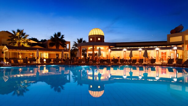 Una piscina con sillas de descanso y sombrillas, un edificio con cúpula y palmeras.