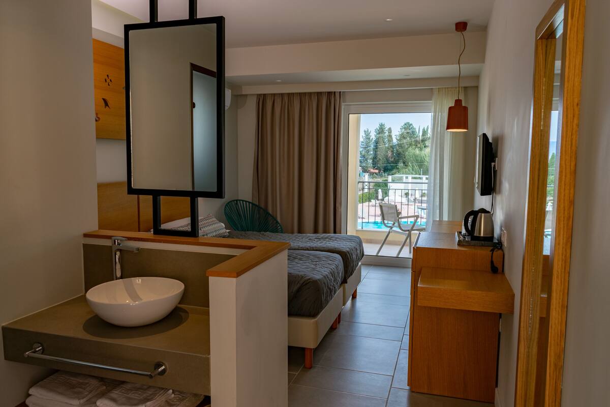 A hotel room with a large bed, a bedside table with a kettle, a bathroom with a sink and mirror, and a view of the outdoors.