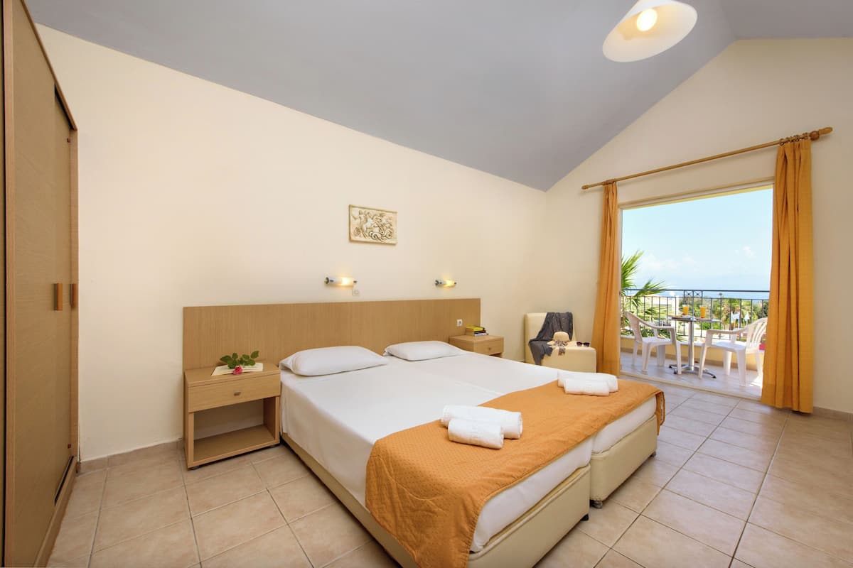 A hotel room with a large bed, a bedside table, a nightstand, a chair, a small table with chairs outside, and a balcony with a view.