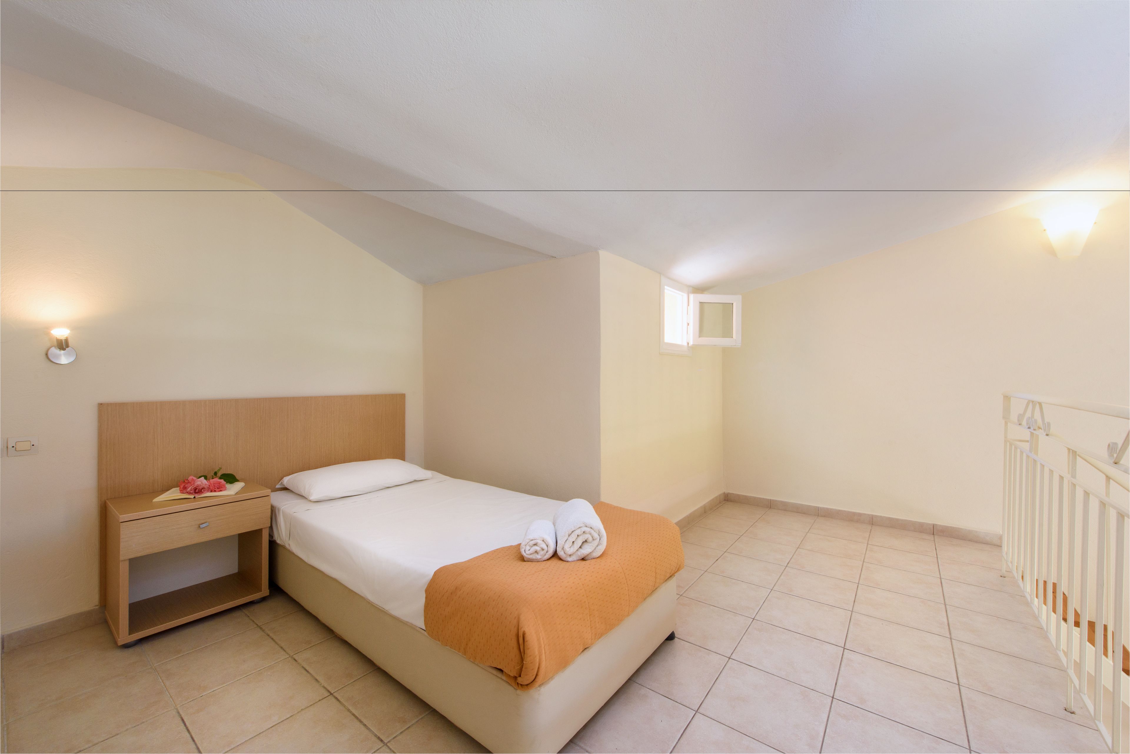 Ein Schlafzimmer mit einem Bett, einem Nachttisch, einem kleinen Fenster und einem weißen Geländer.