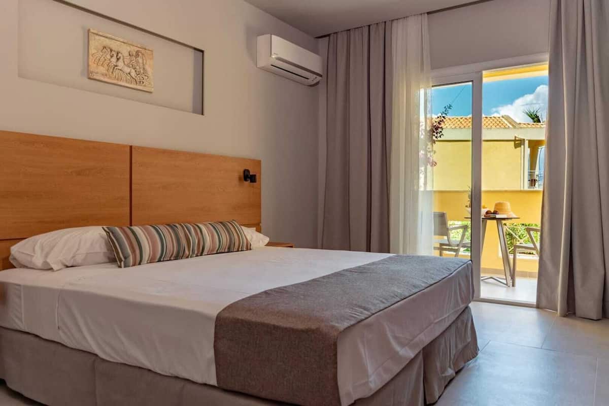 A hotel room with a large bed, a wooden headboard, a wall-mounted air conditioner, and a view of the outside balcony.