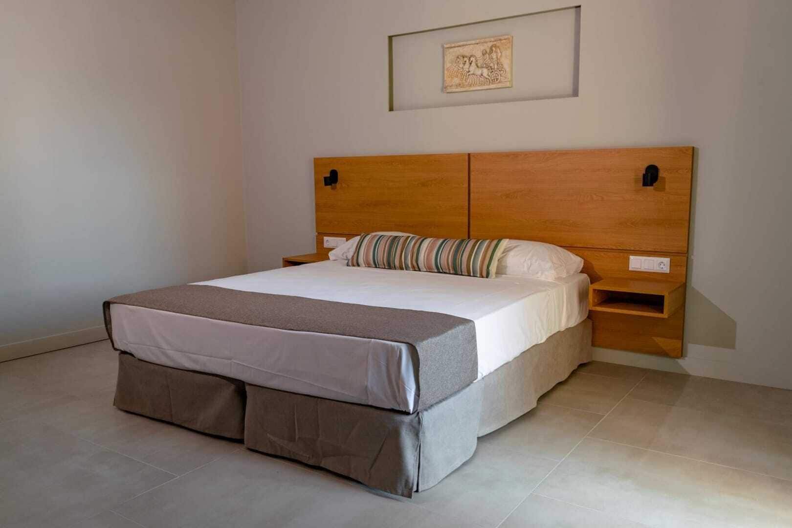 Een opgemaakt bed met een houten hoofdbord, een nachtkastje en een klein wandrek.