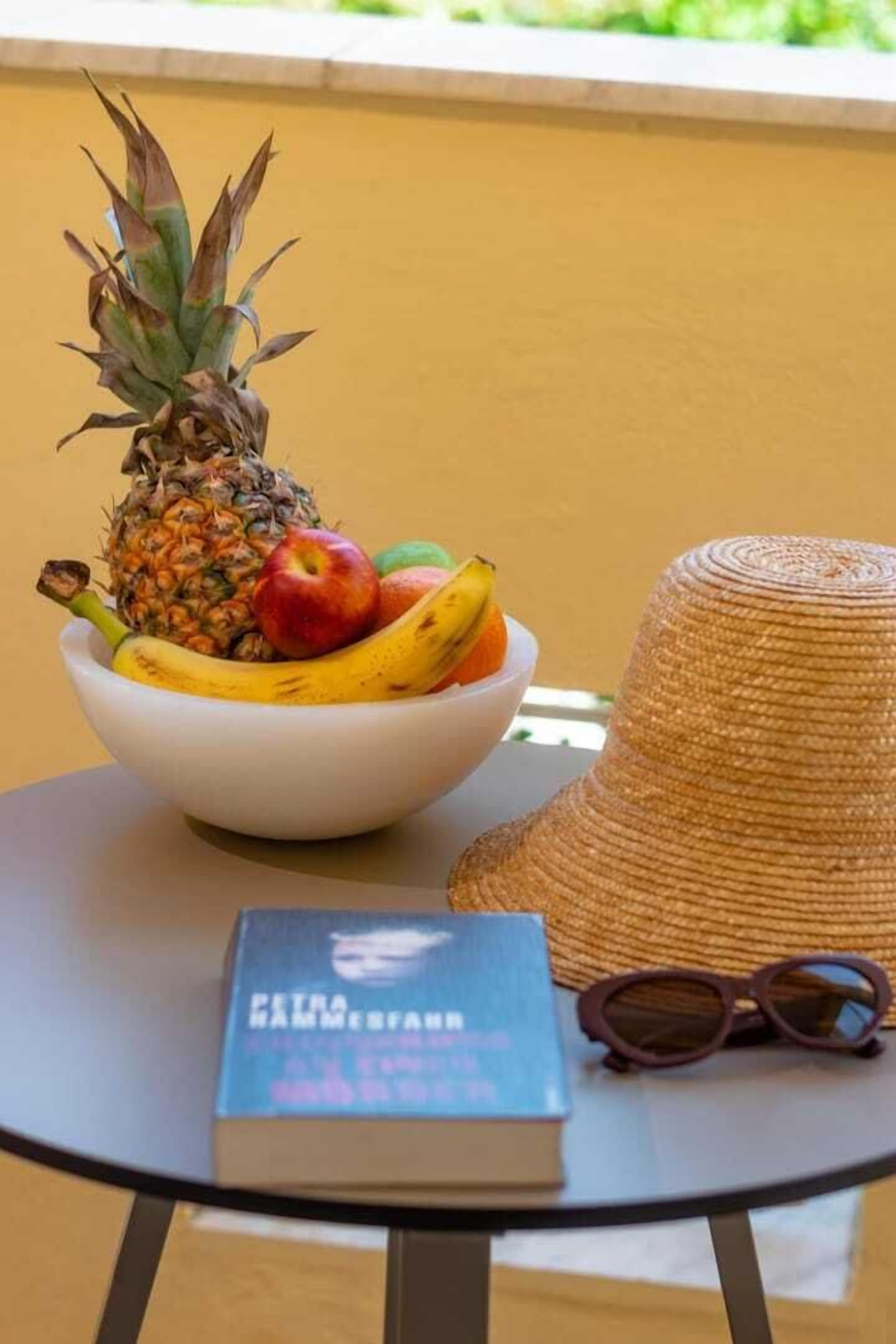 Un saladier rempli de fruits est posé sur une table, à côté d’un chapeau de paille et d’une paire de lunettes de soleil.