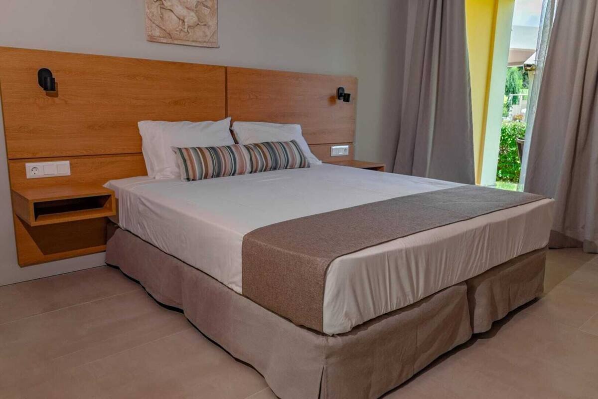 A neatly made bed with a wooden headboard, a bedside table, and a view of greenery outside.