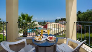 Un balcón con dos sillas blancas, una mesa redonda con platos de desayuno, y vistas a una piscina y edificios al fondo.