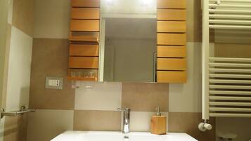 Un baño con lavamanos blanco, tocador de madera y espejo con persianas de madera. Hay plantas en macetas en el alféizar de la ventana y un perchero a la derecha.