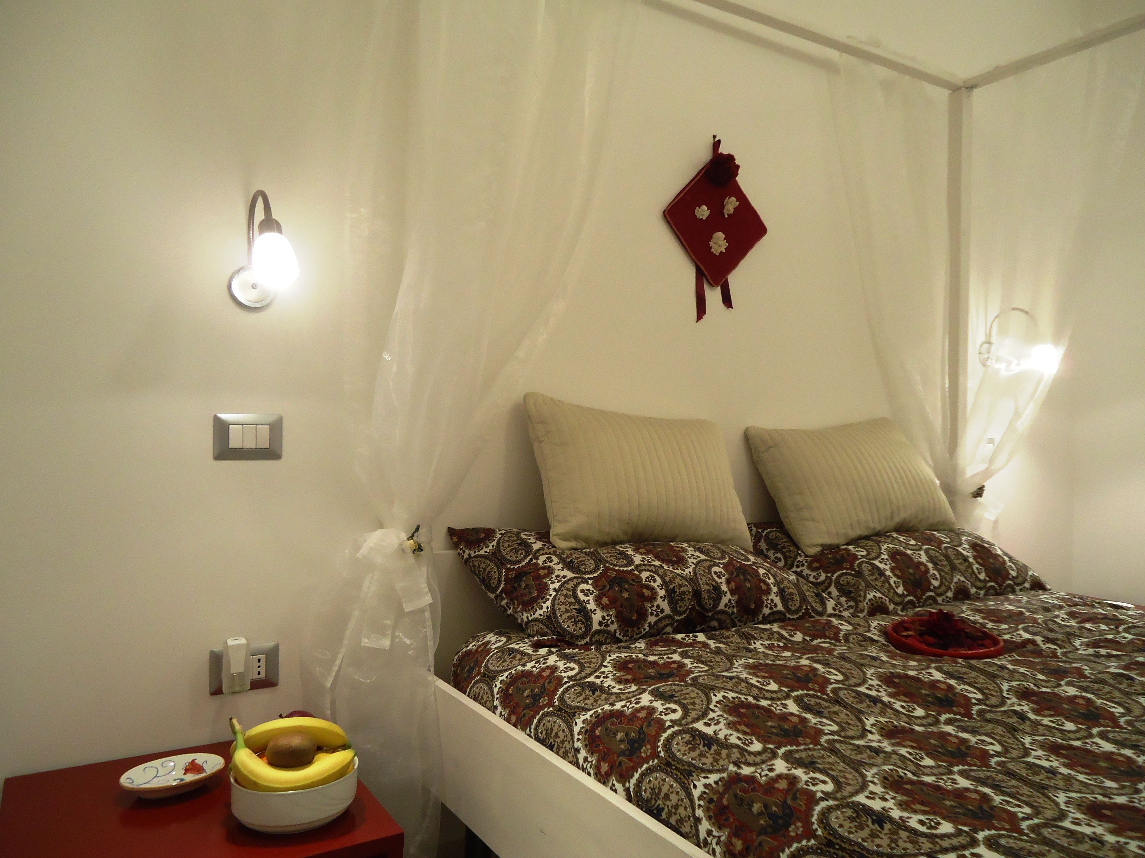 Une chambre à coucher avec un lit, une table de chevet sur laquelle se trouve un bol de fruits, une lampe fixée au mur et une prise de courant.