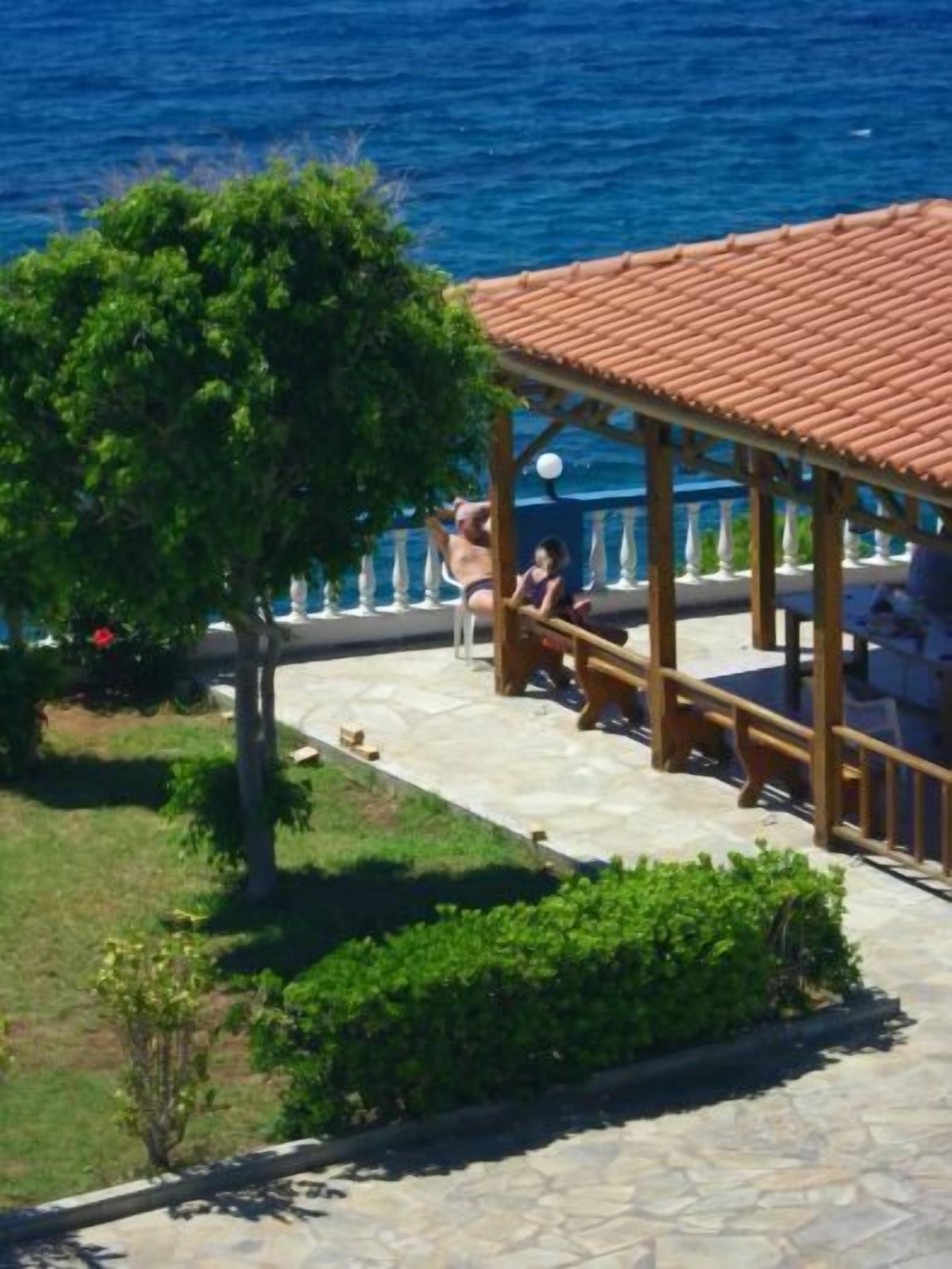 A covered outdoor seating area with a tiled roof, surrounded by greenery and overlooking a body of water.