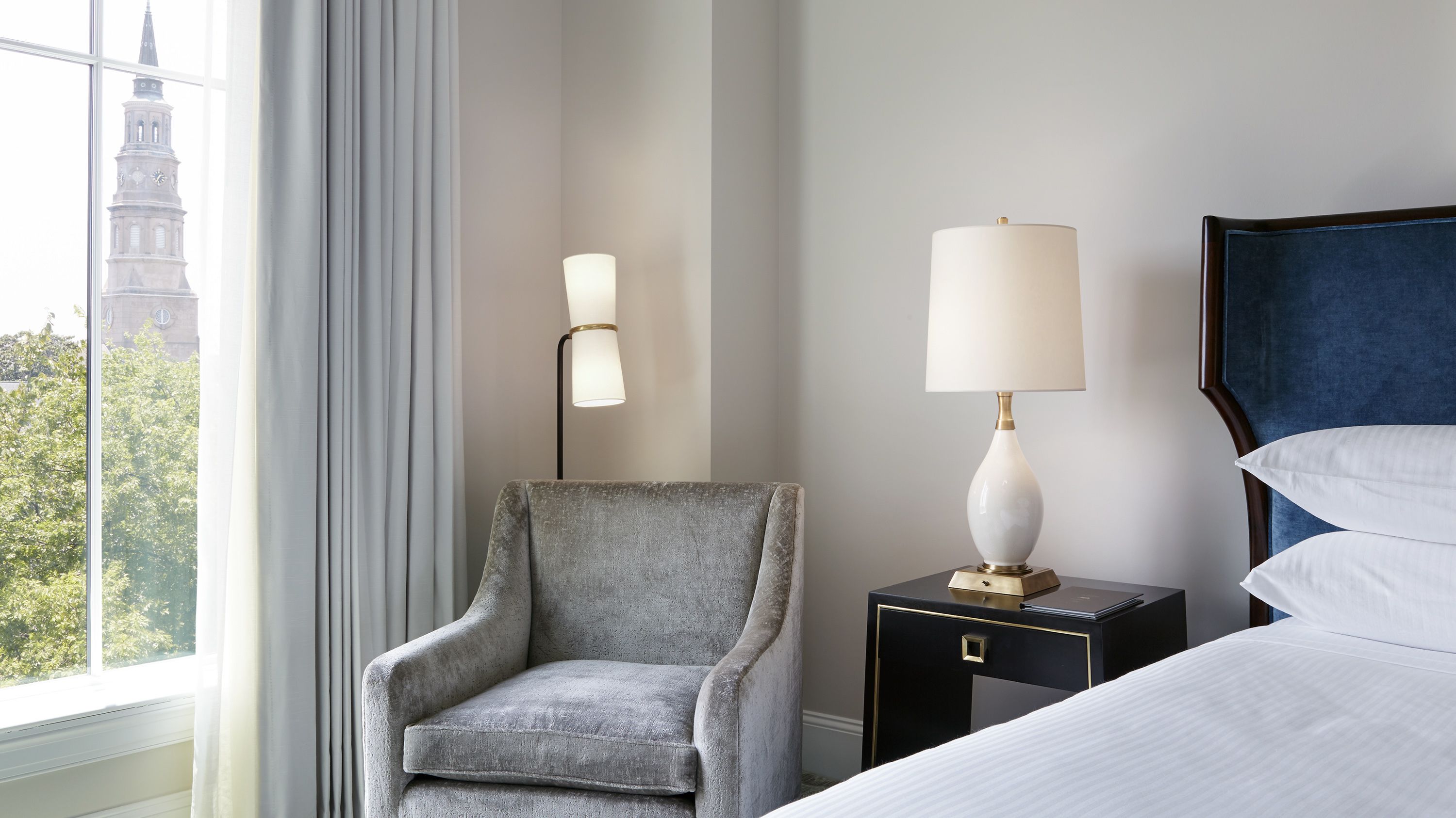 A hotel room with a grey armchair, a bedside table with a lamp, a bed with white linens, and a view of a building through a window.