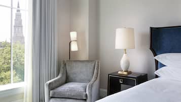 A hotel room with a grey armchair, a bedside table with a lamp, a bed with white linens, and a view of a building through a window.