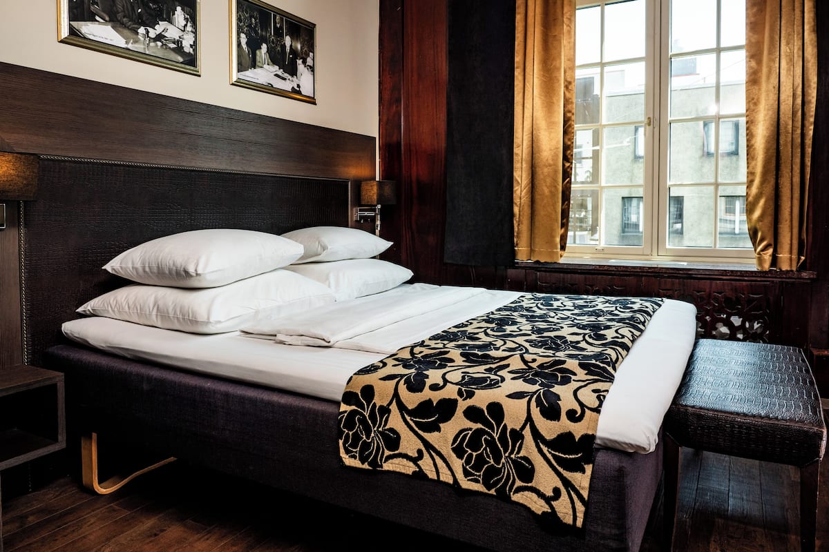 A neatly made bed with white linens and a floral-patterned throw, a wooden headboard, and a window with curtains.