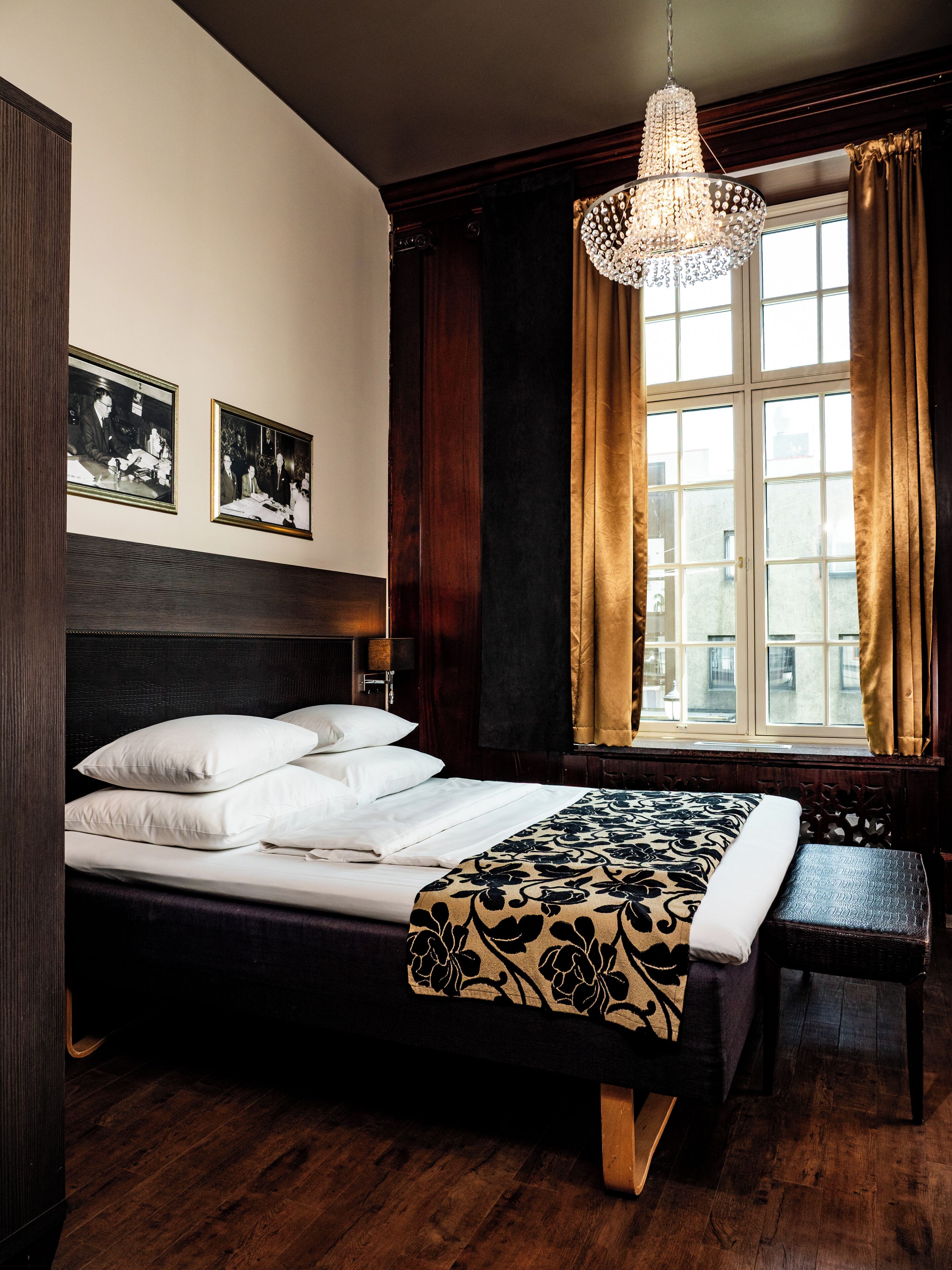 Una habitación con cama, un banco, una araña y dos cuadros en la pared.