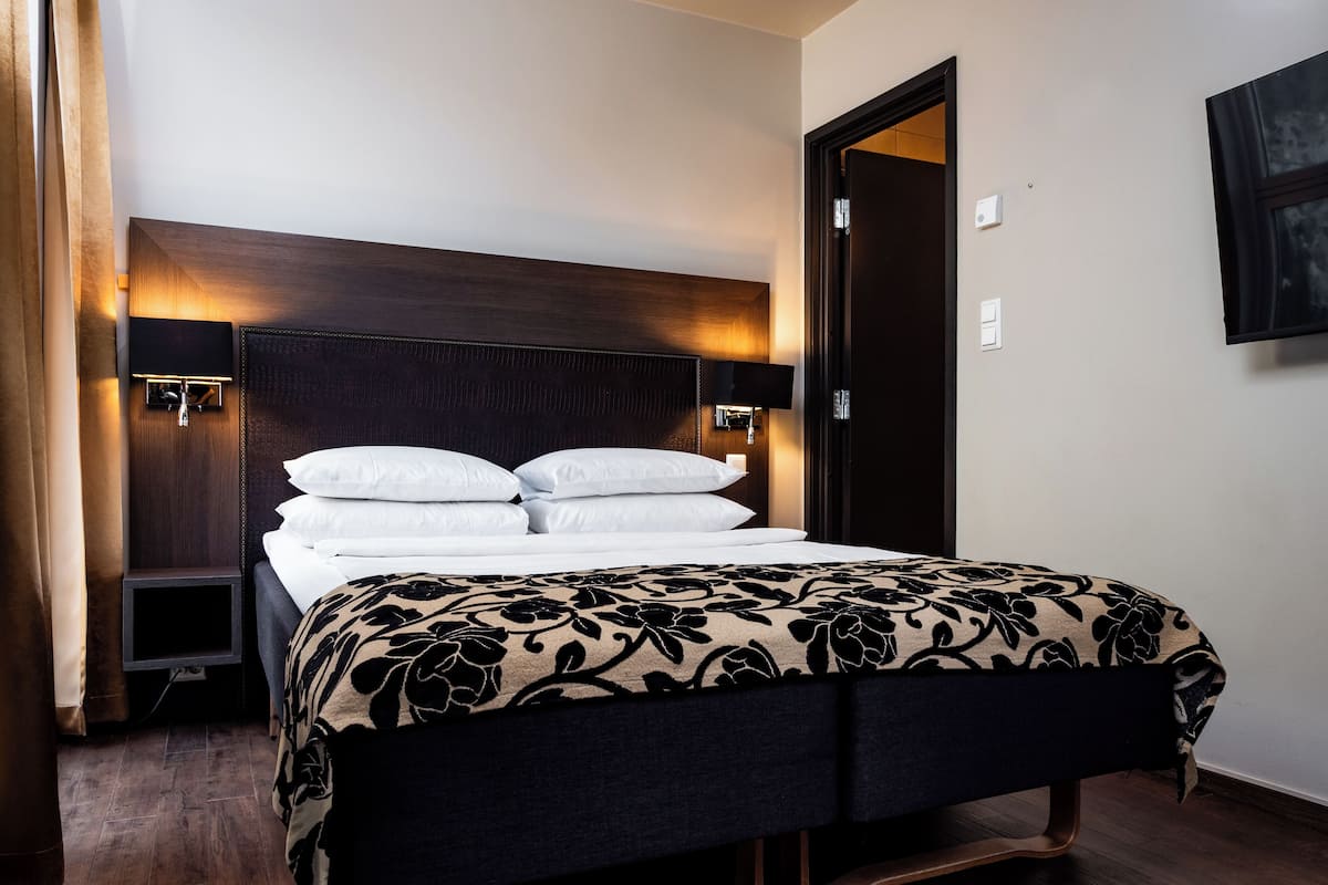 A neatly made bed with a dark headboard and bedside lamps, a wooden floor, and a door to the right.