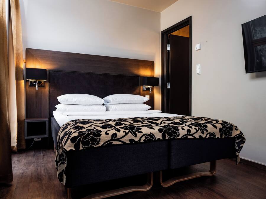 A neatly made bed with a dark headboard and bedside lamps, a wooden floor, and a door to the right.