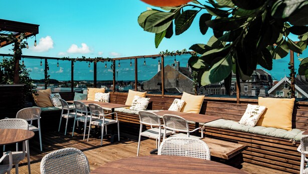 An outdoor seating area with wooden tables and chairs, cushioned benches, and a view of the cityscape. - Banken Hotel (Haugesund)