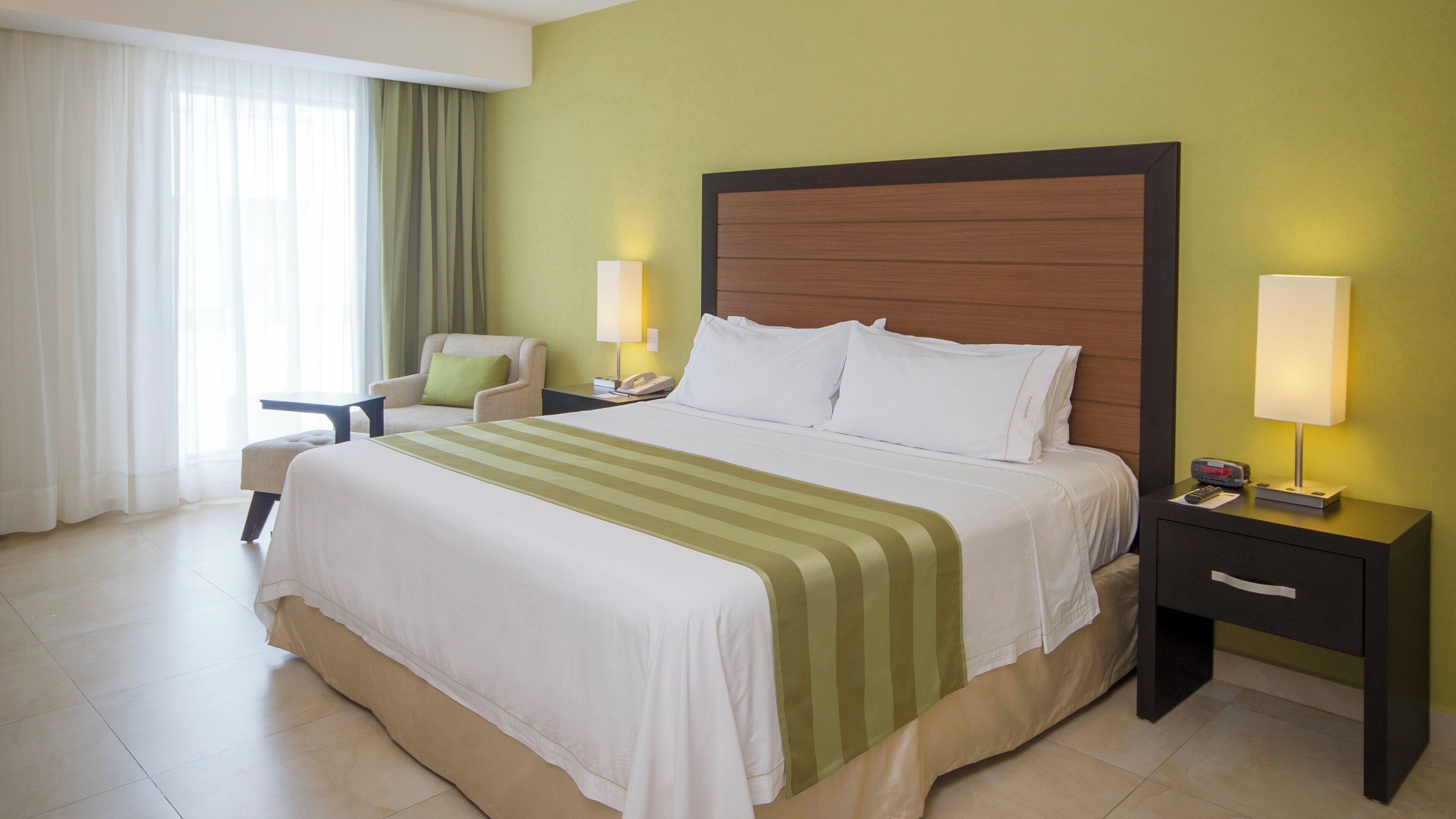 A hotel room with a large bed, a chair, a nightstand with a lamp, and a bedside table with a clock.