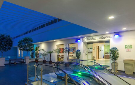 An indoor hotel lobby with an escalator, potted plants, and a Holiday Inn Express sign.