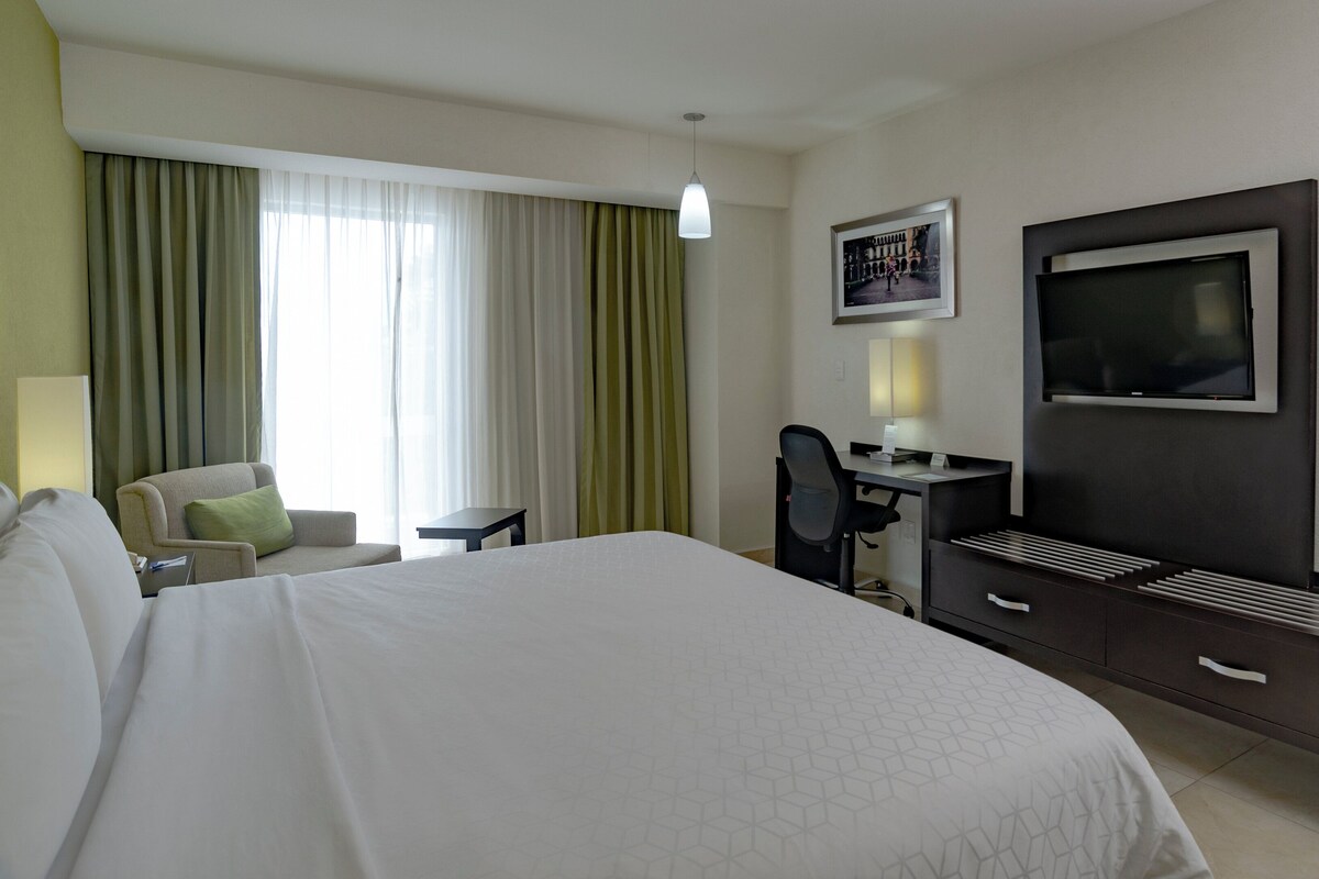 A hotel room with a large bed, a desk with a chair, a television, and a window with curtains.