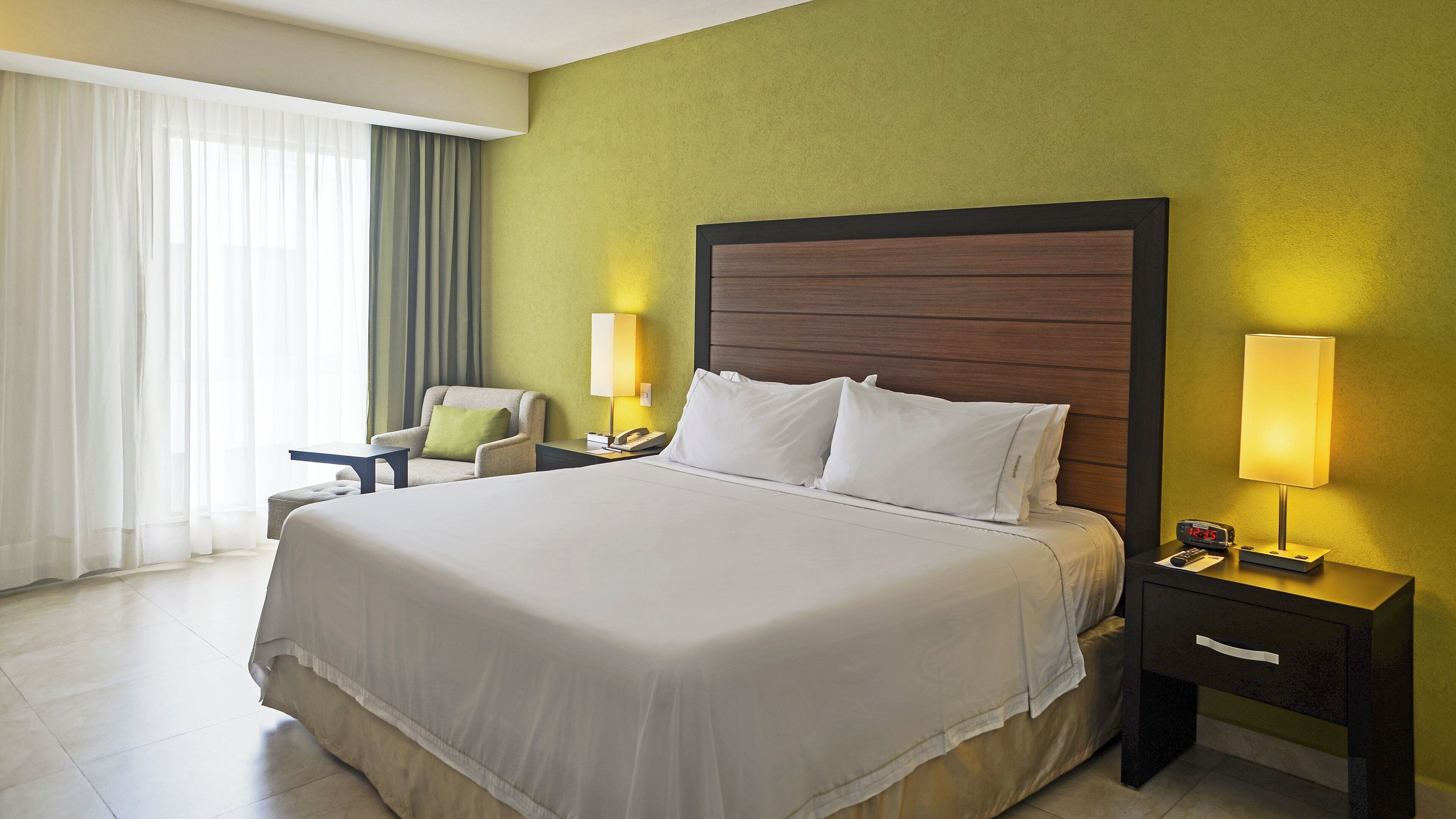 A hotel room with a large bed, a chair, a nightstand with a lamp, and a wall clock.