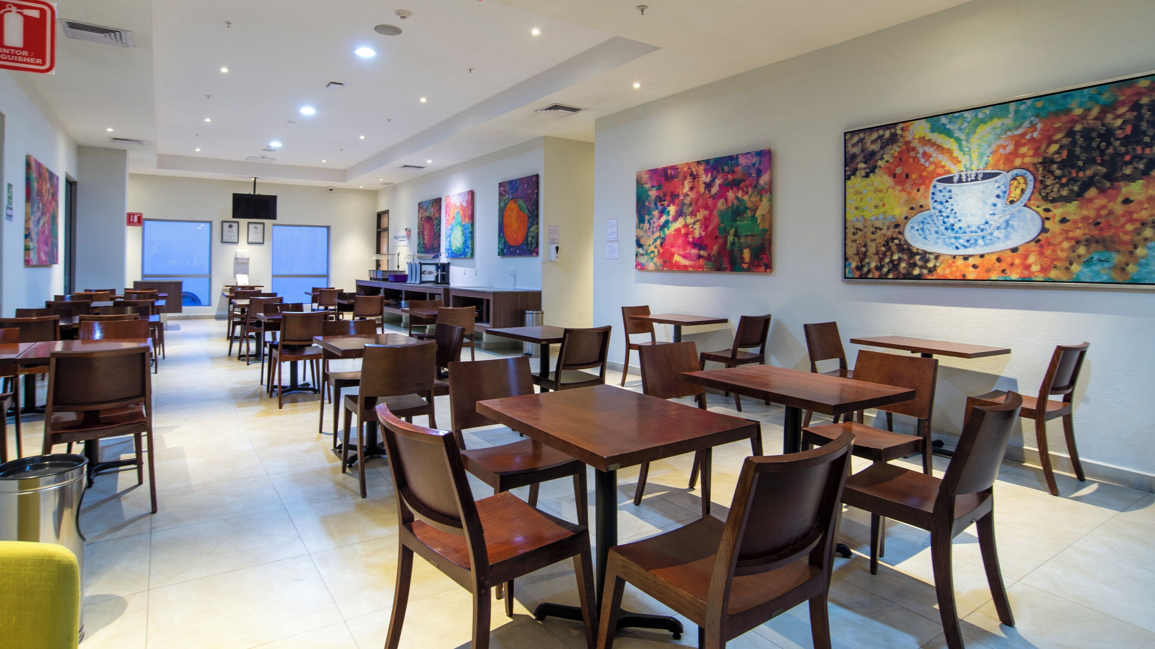 A spacious dining area with multiple wooden tables and chairs, a fire extinguisher, and colorful abstract paintings on the walls.