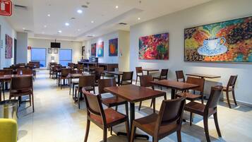 A spacious dining area with multiple wooden tables and chairs, a fire extinguisher, and colorful abstract paintings on the walls.