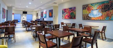 Un comedor amplio con varias mesas y sillas de madera, un extintor, y pinturas abstractas de colores en las paredes.