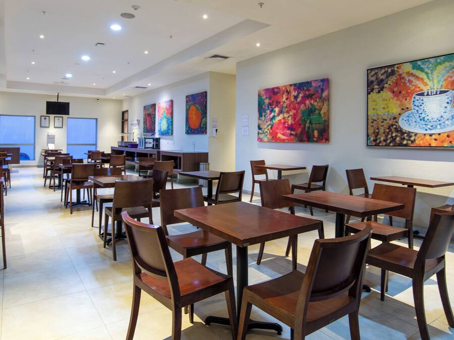 A spacious dining area with multiple wooden tables and chairs, a fire extinguisher, and colorful abstract paintings on the walls.