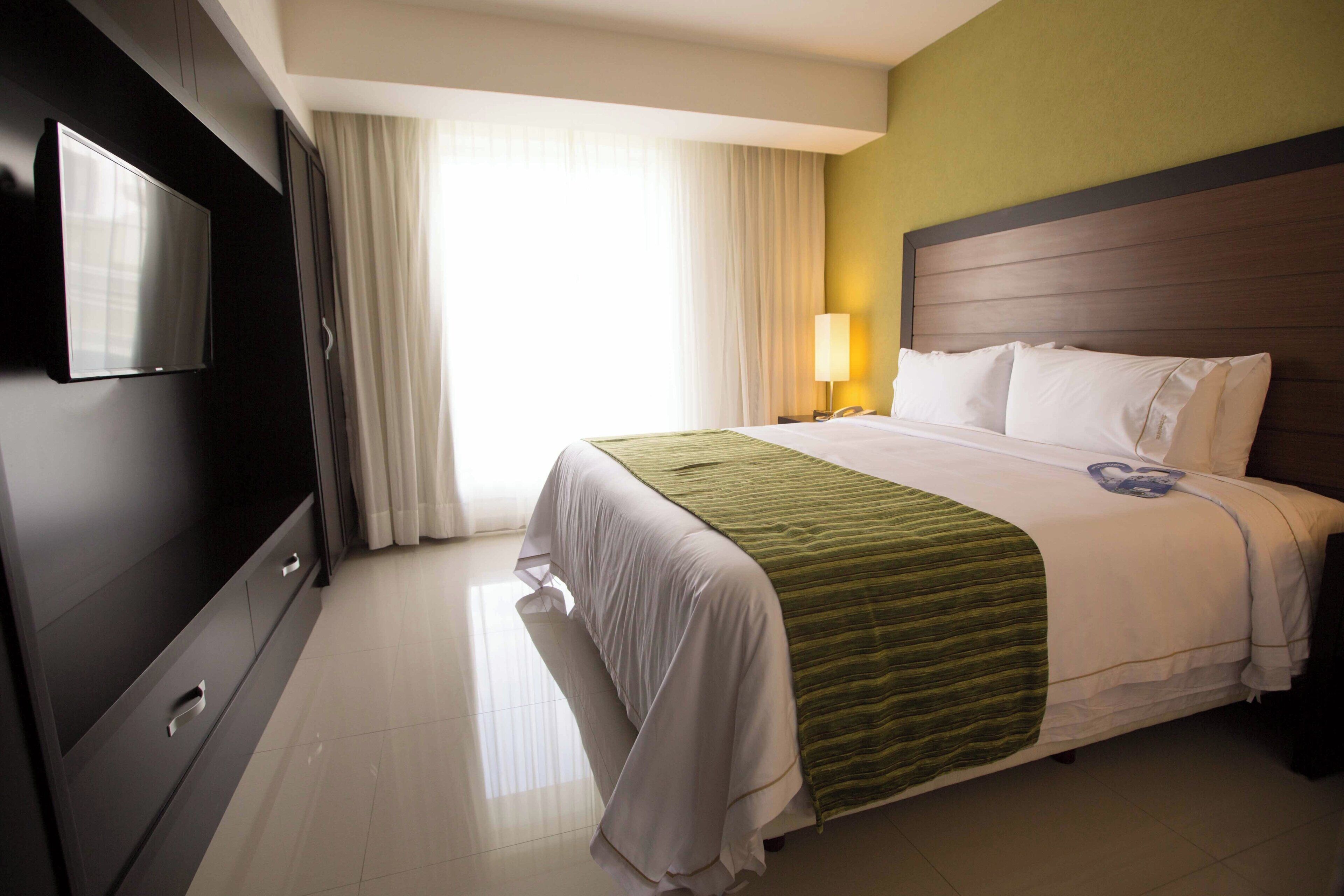 A hotel room with a bed, a TV, a lamp, and a window with curtains.