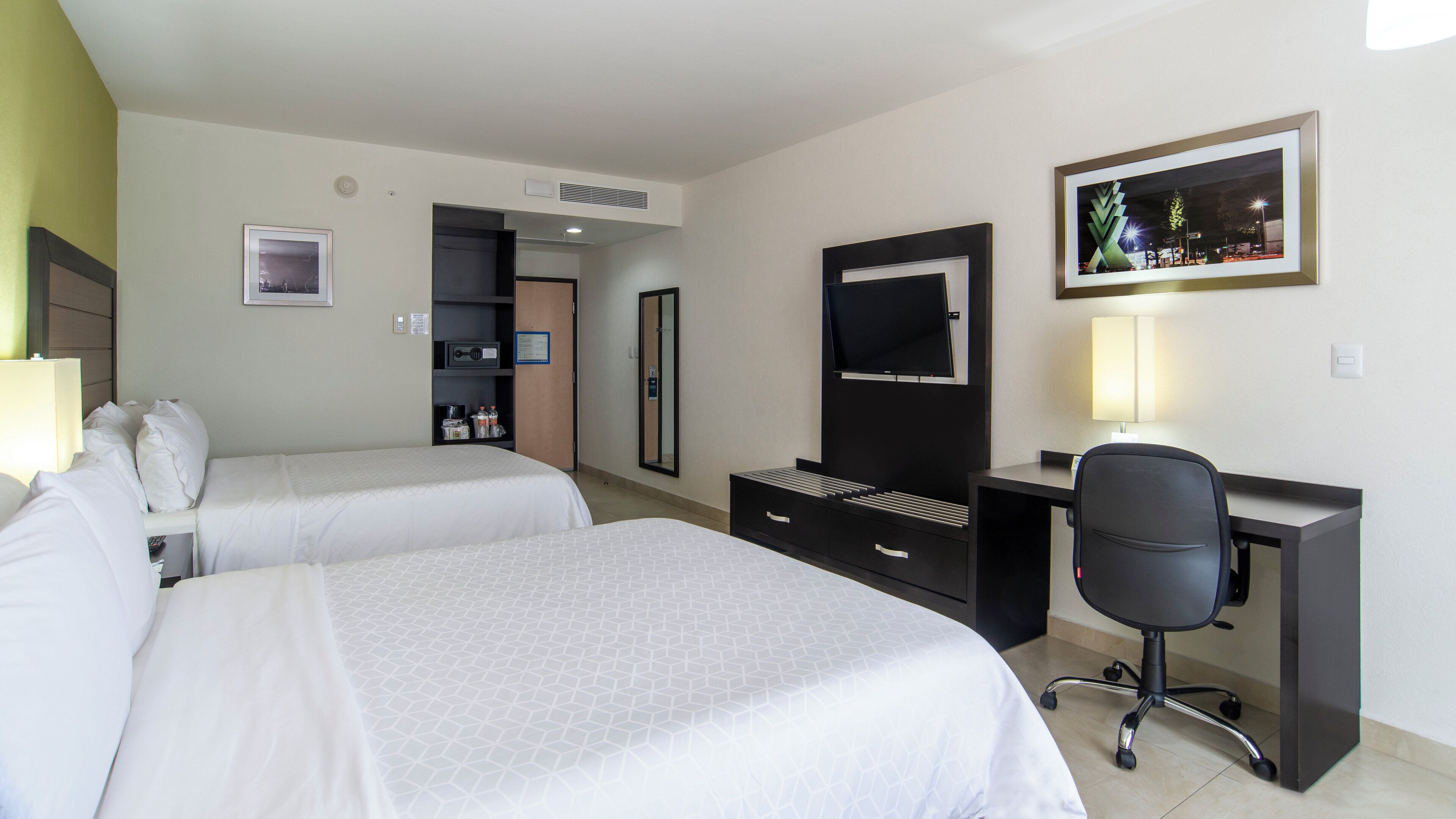 Habitación de hotel con dos camas, un escritorio, una silla, un espejo, un televisor y un cuadro en la pared.