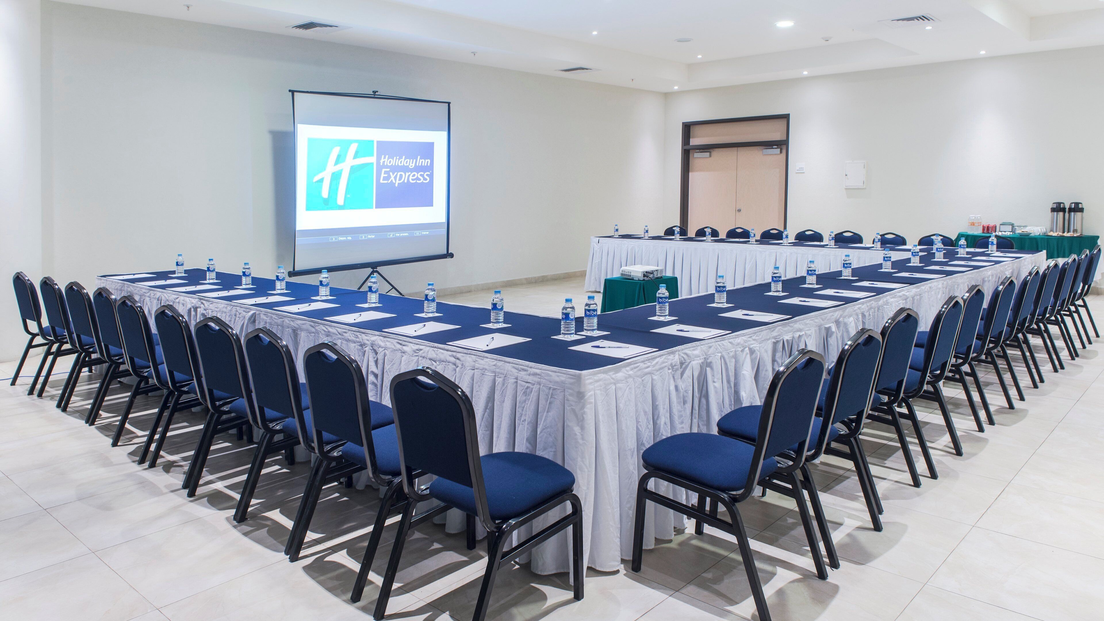 Una sala de conferencias con una larga mesa, sillas, una pantalla de proyector y el logotipo de Holiday Inn Express.