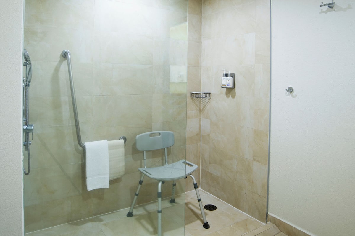 A shower area with a glass door, a handheld showerhead, a grab bar, and a small step stool.
