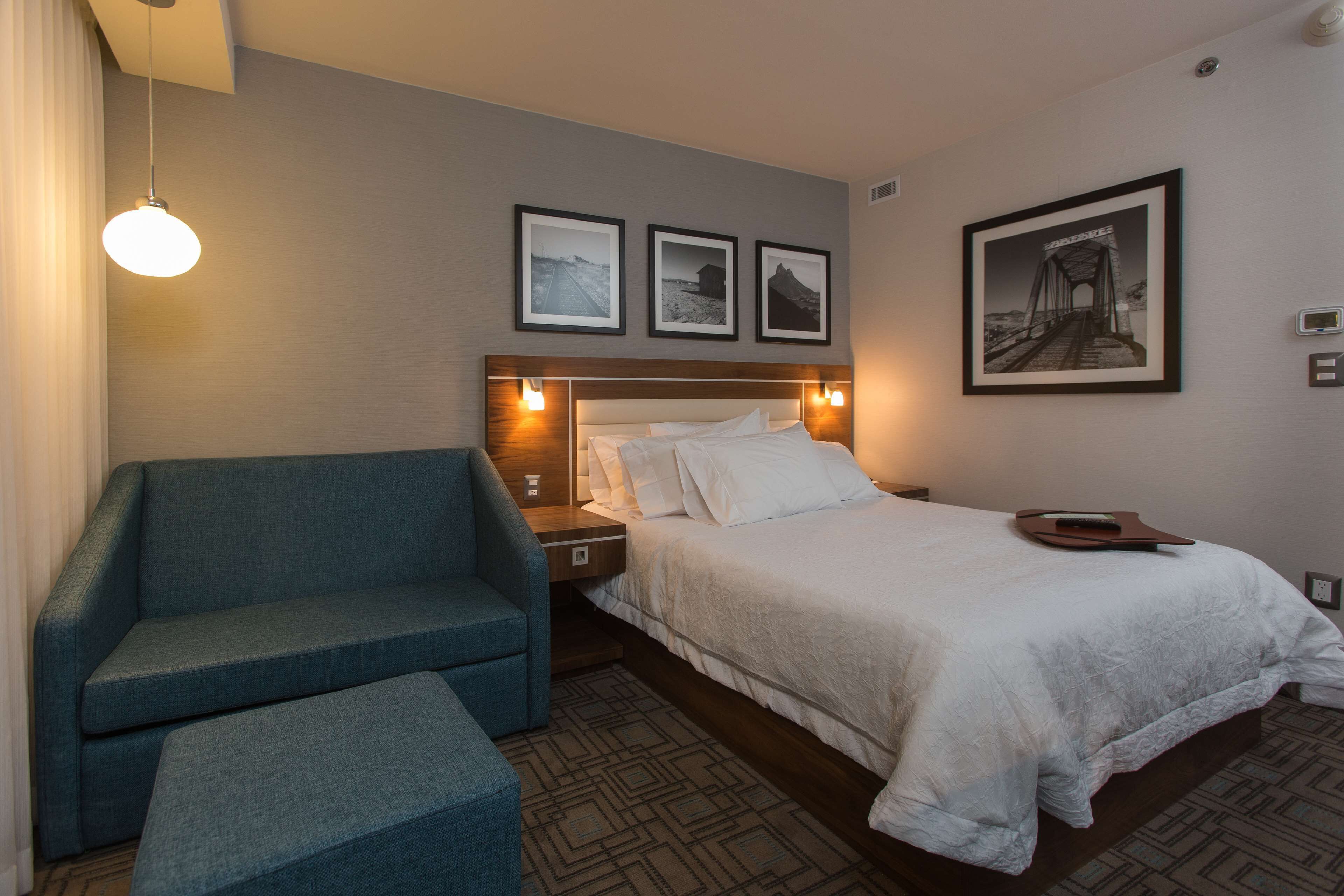 Habitación de hotel con una cama, un sillón azul, una mesita de noche y un cuadro colgado en la pared que muestra un puente.