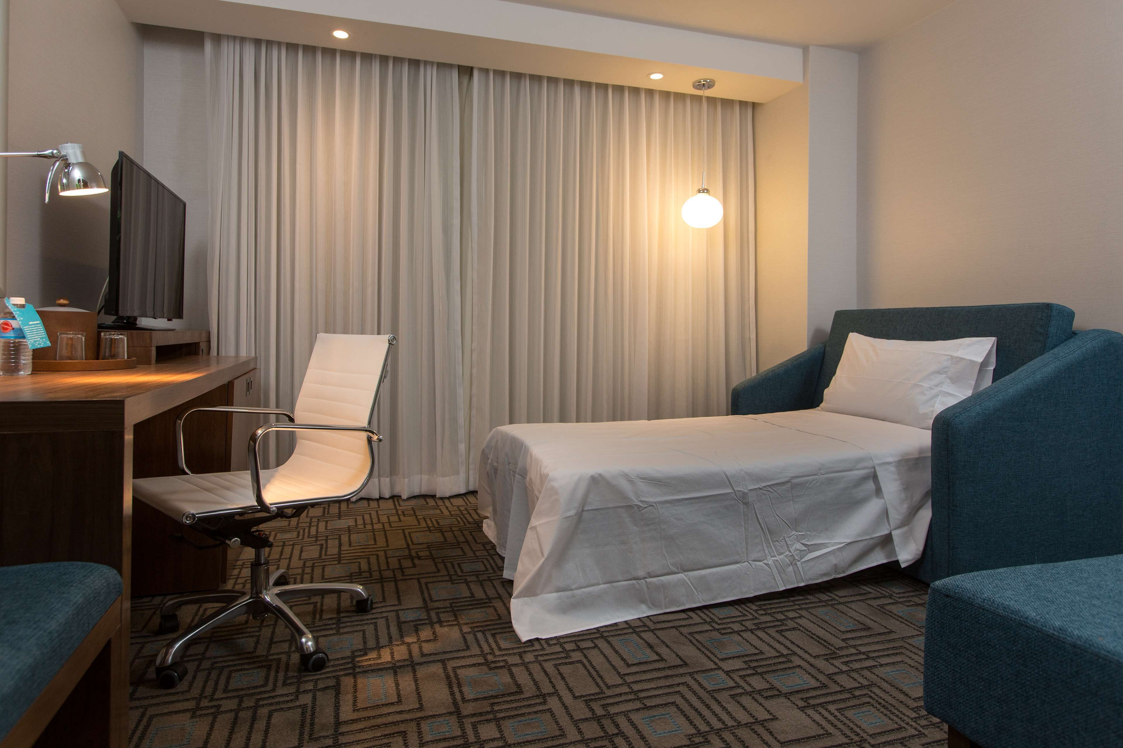 Habitación de hotel con cama, dos sillones, escritorio con computadora y lámpara, y televisión.