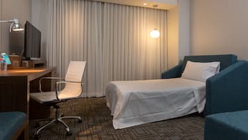 Habitación de hotel con cama, dos sillones, escritorio con computadora y lámpara, y televisión.