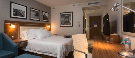 A hotel room with a bed, a desk, a chair, a TV, and framed pictures on the wall.