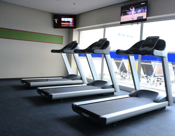 Un gimnasio con cintas de correr, un televisor mostrando un programa y un ventanal con vista a una estructura azul en el exterior.