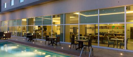 A swimming pool with a sunken seating area, surrounded by a building with large windows and a dining area visible inside.
