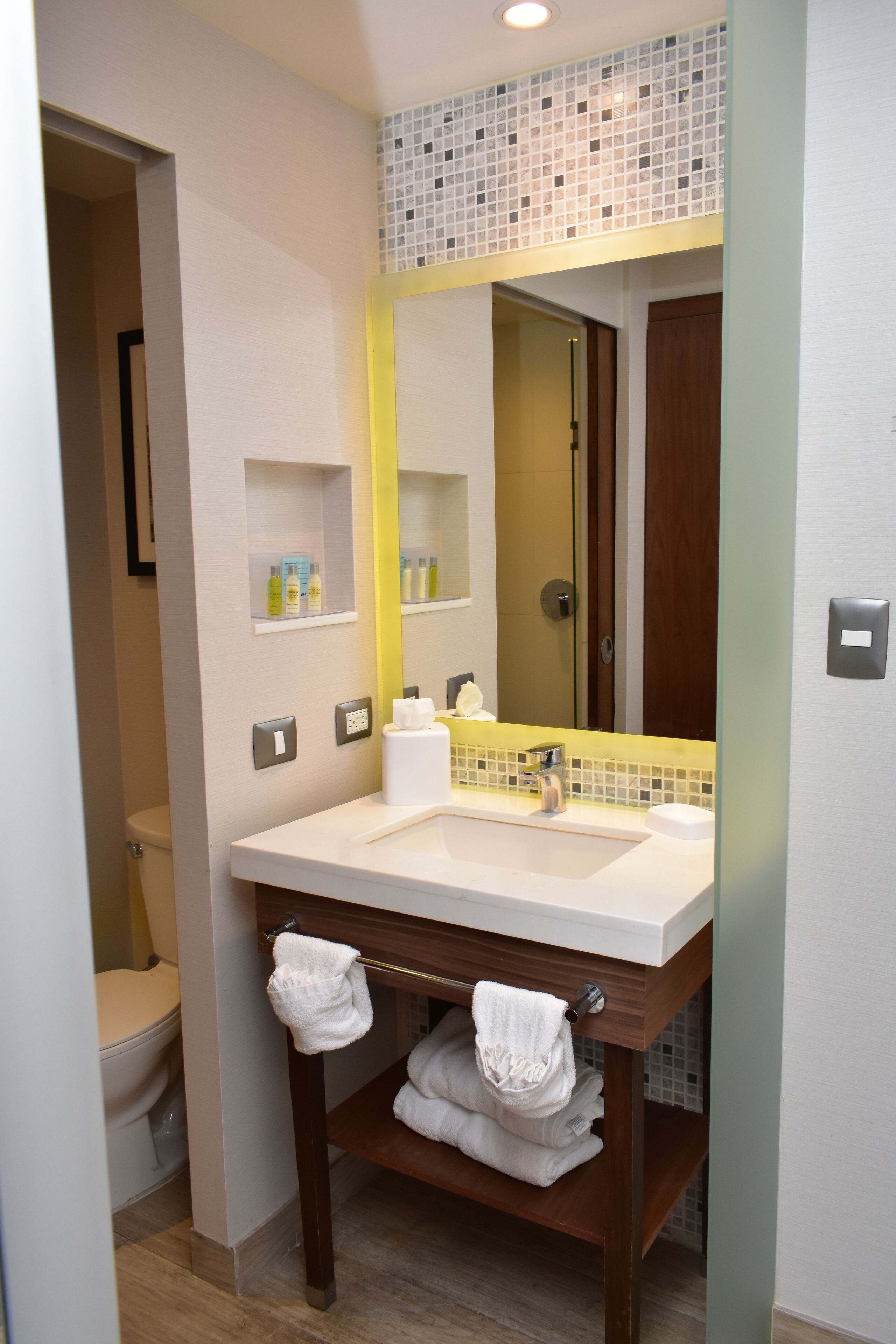 A modern bathroom with a large mirror, a sink, and a toilet. There are shelves with toiletries and a towel rack with towels.