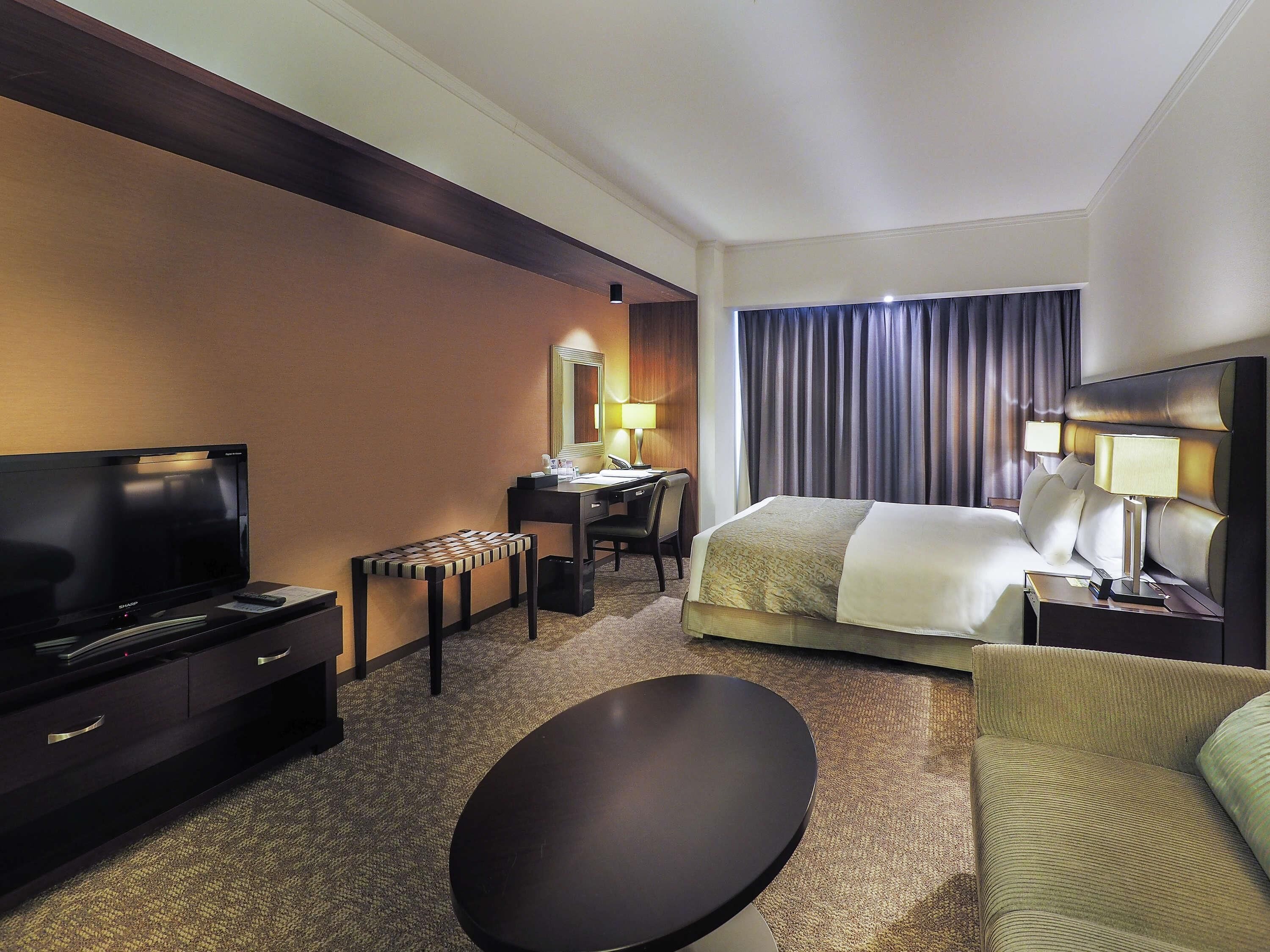 A hotel room with a bed, bedside tables, a desk with a chair, a television, and a small table.