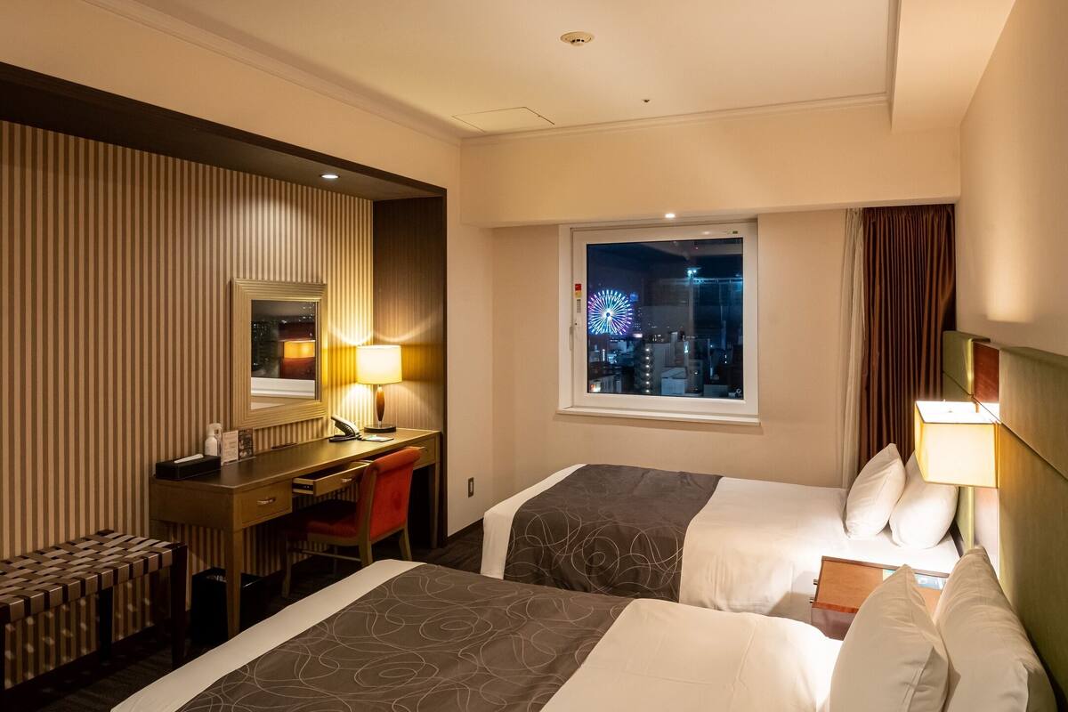 Habitación de hotel con dos camas, un escritorio y una silla, un espejo, una ventana con vistas a la ciudad y un televisor.