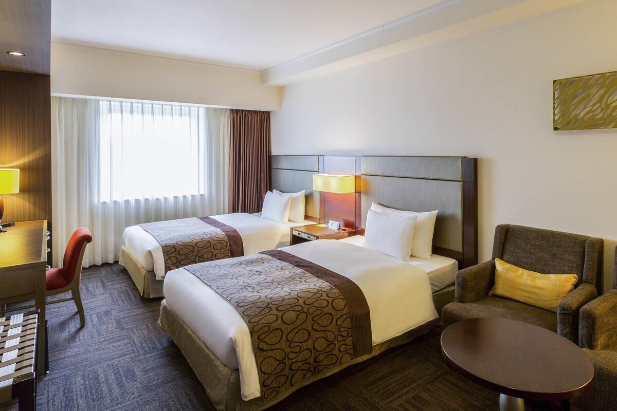 A hotel room with two beds, a desk, a chair, a small table, and a window with curtains.