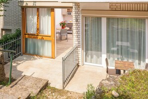 Patio esterno moderno con tavolo e sedie in legno, un piccolo giardino e una porta scorrevole in vetro che dà su un ambiente interno con cucina e sala da pranzo.